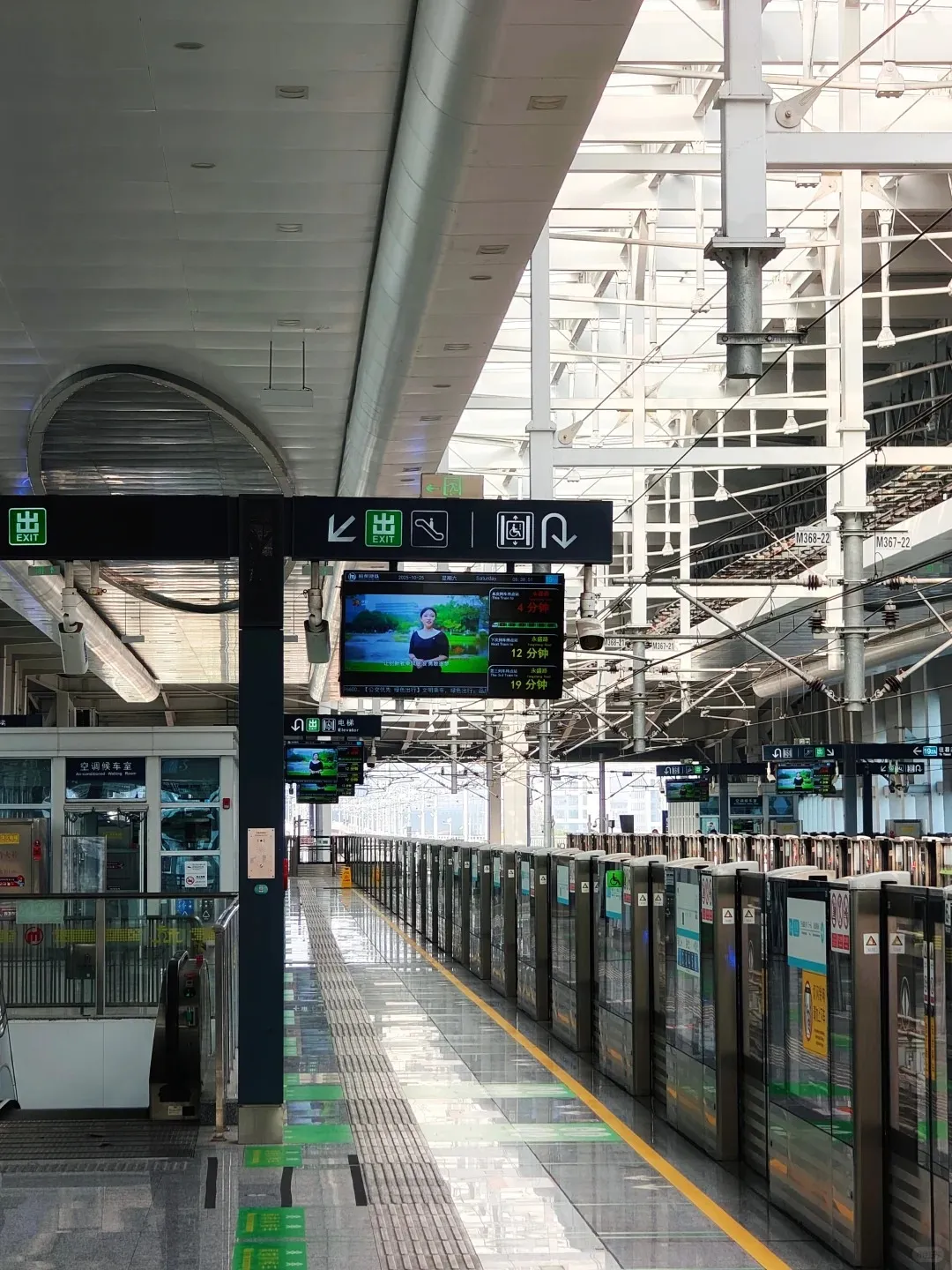 An empty, modern Hangzhou metro station platform with grey tiled flooring and yellow tactile paving. A long line of closed glass platform screen doors stretches along the track. Overhead, multiple digital displays show advertisements and train departure times in Chinese characters. Signs for 'EXIT', 'ELEVATOR', and 'ESCALATOR' are visible on black panels. On the left, a glass-walled 'Air-conditioned Waiting Room' is visible. The station features a high, light-filled ceiling with exposed white structural beams and overhead power lines in the background.