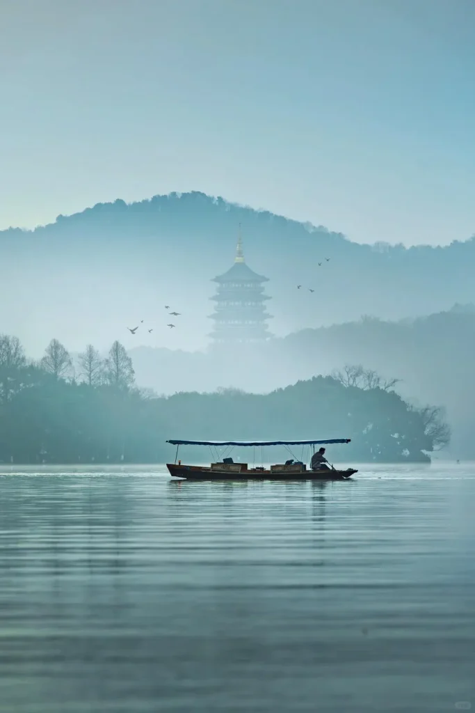 A person rows a traditional wooden boat with a blue canopy on a calm, misty lake. In the background, a multi-tiered pagoda with a golden spire is partially obscured by fog, surrounded by hazy mountains and trees, with several birds flying overhead.