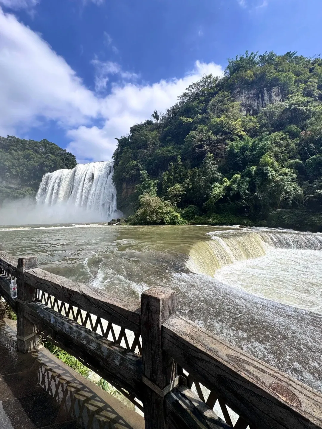 The massive Huangguoshu Waterfall cascades from a lush green karst mountain on the left, under a blue sky with white clouds. In the foreground, a brown wooden railing overlooks a wide, turbulent river with a smaller, wide waterfall.