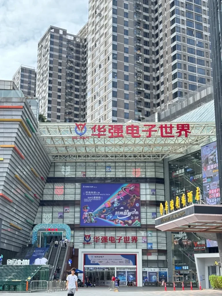 Exterior view of the Huaqiang Electronic World (HQ-Mart) building in Shenzhen, a multi-story modern complex featuring red Chinese characters, a large LED screen advertising gadgets and tech fashion, escalators leading to upper floors, and pedestrians near the entrance, with several tall residential buildings visible behind it.