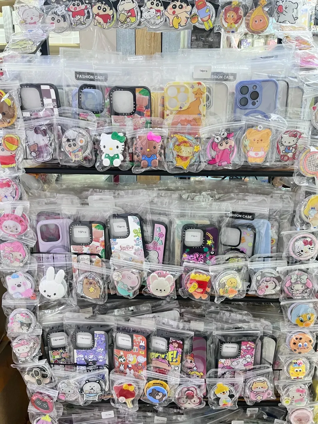 A retail display wall packed with hundreds of individually packaged smartphone cases and phone grips. The accessories showcase a diverse array of colorful designs, including popular cartoon characters like Hello Kitty and Crayon Shin-chan, animals, and food items. Labels indicating iPhone models such as '15 Pro' and '14 Pro Max' are visible on some cases.