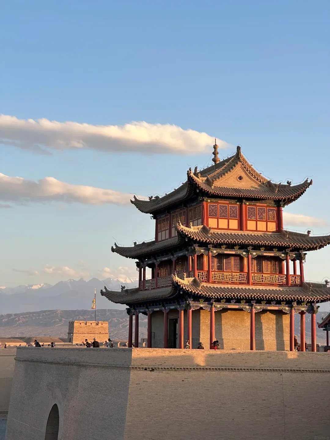 A multi-tiered traditional Chinese fortress tower, with red pillars, intricately patterned windows, and dark tiled roofs, stands prominently on a wide, light-brown brick wall. Distant snow-capped mountains are visible under a blue sky with scattered clouds. The scene is illuminated by warm, golden sunlight.