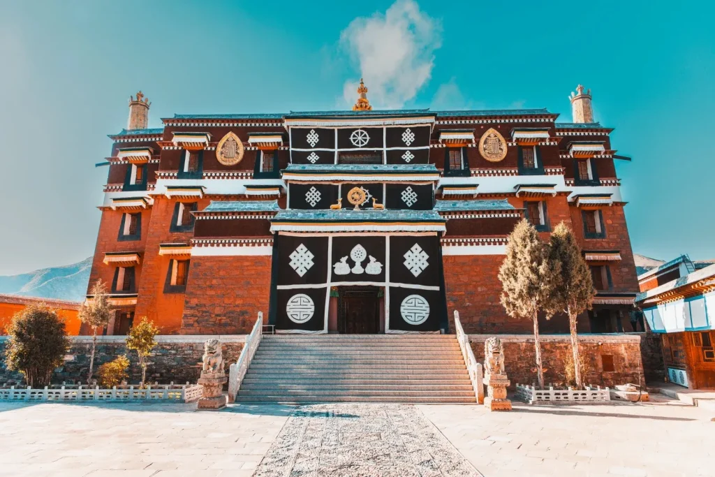 A wide, frontal view of Labrang Monastery's Great Sutra Hall, a multi-story building with reddish-brown stone walls. Its central section features dark fabric hangings adorned with white auspicious Buddhist symbols and a golden Dharma wheel flanked by two deer above the main entrance. A broad stone staircase, guarded by stone lions, leads to the entrance. The building is set against a bright blue sky.