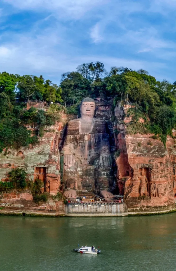 A colossal statue of the seated Buddha carved into a red sandstone cliff face, overlooking a wide, green river. The statue is immense, with visible details of its serene face, folded hands, and large feet resting on a platform. Lush green trees and foliage grow along the top and sides of the cliff. A small white boat with a few people on board is visible in the river in the foreground.