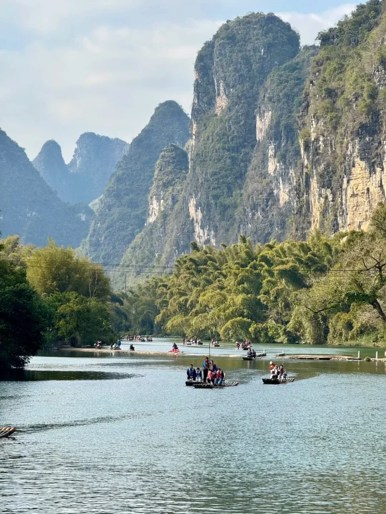 Several motorized bamboo rafts carrying tourists float on a calm river. The riverbanks are lined with dense green bamboo forests, and in the background, numerous towering grey-green karst mountains rise against a bright, hazy sky.