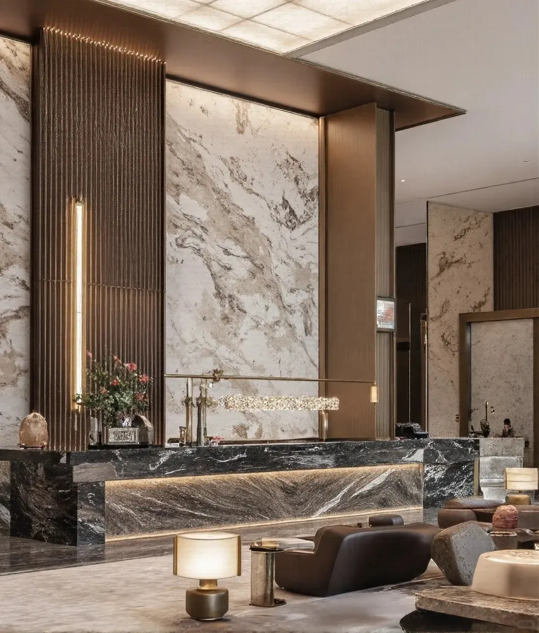 A luxurious, modern hotel reception desk made of dark, veined marble with warm underlighting. Behind it, a wall of white and grey marble is flanked by tall, fluted bronze panels with a vertical LED light. A long, sparkling chandelier hangs above the desk. In the foreground, a stylish lounge area features dark, abstract seating and a contemporary table lamp.