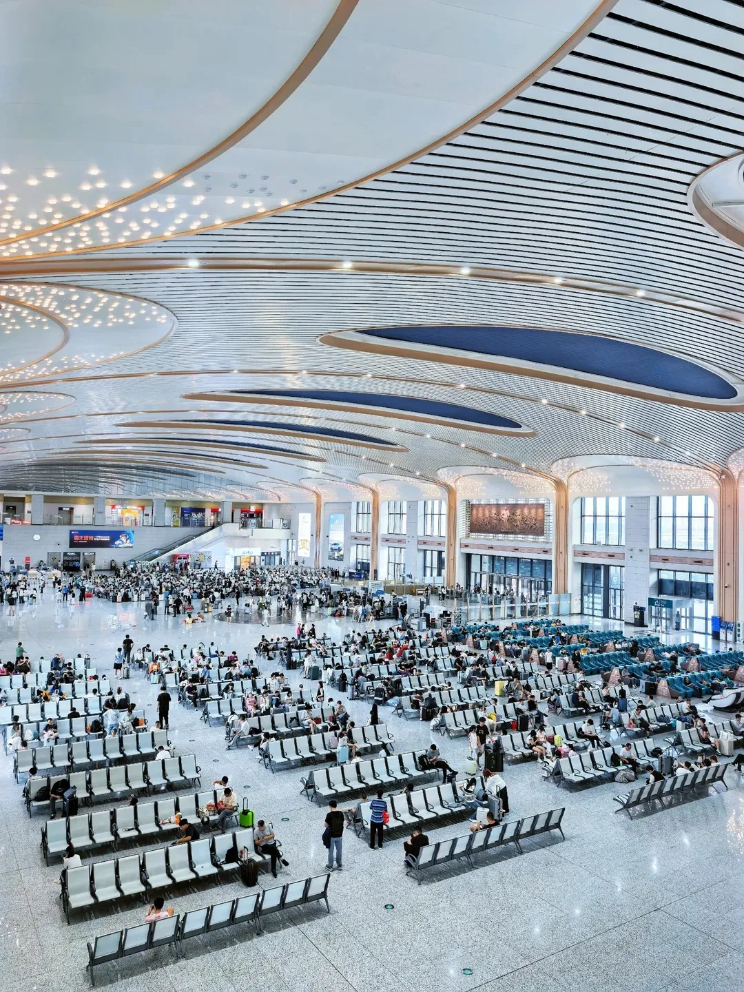A vast, modern railway station departure hall with a high, elaborately designed ceiling featuring illuminated circular patterns and layered metallic louvers. Rows of grey, bench-style seating are arranged on a polished floor, filled with numerous people, including families with children, waiting with luggage. Large windows and high ceilings create a bright and spacious atmosphere. Escalators and signs are visible in the background.