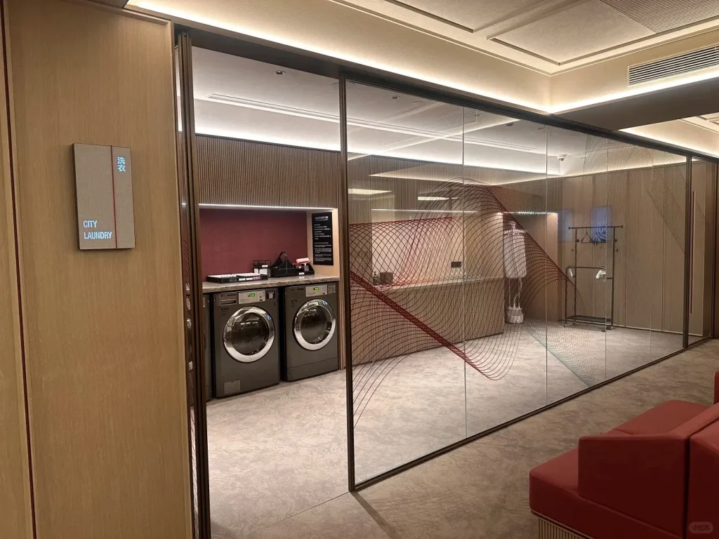 A modern hotel self-service laundry room in China, featuring two front-loading washing machines with chrome doors, a dark red tiled backsplash, and light wood cabinetry. A sign on a wooden wall to the left reads 'CITY LAUNDRY' and '洗衣'. The laundry area is enclosed by glass walls with decorative red and green wavy patterns, and a red upholstered waiting bench is visible in the foreground.