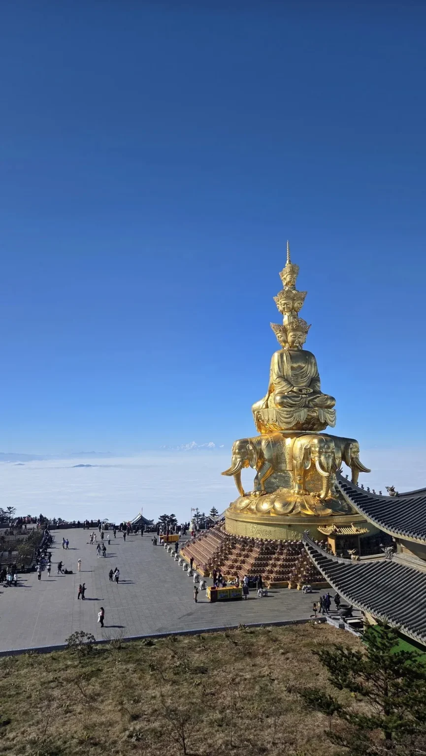 A large, ornate, golden Buddhist statue with multiple heads and faces sits atop a multi-tiered pedestal supported by elephants. The statue is situated on a mountain summit above a vast expanse of white clouds, with a clear blue sky above. In the foreground and to the left, a paved plaza is filled with numerous tourists. To the right, traditional Chinese temple roofs are visible.