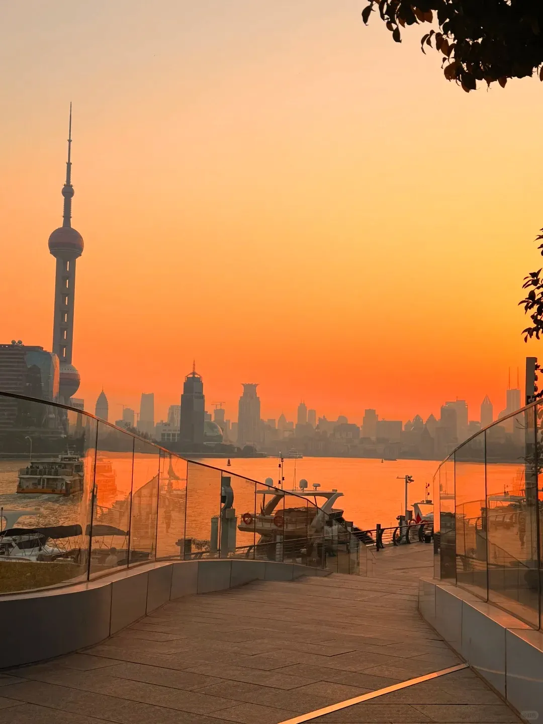 A modern paved waterfront path with glass railings in the foreground, leading down towards a river with several boats docked. Across the wide Huangpu River, the Shanghai Pudong skyline is silhouetted against a vibrant orange and yellow sunset sky, with the distinctive Oriental Pearl TV Tower prominently visible on the left.