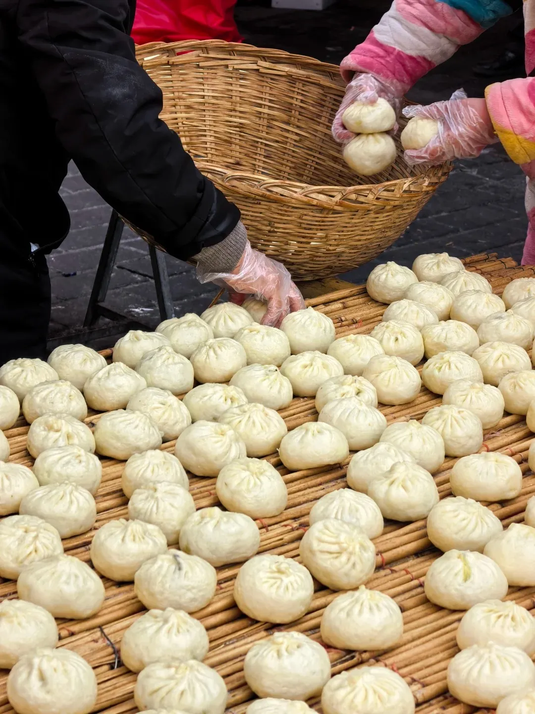 Two individuals wearing clear plastic gloves arrange dozens of freshly prepared white, round, pleated steamed buns on a light brown slatted mat. More buns are visible inside a large woven basket.