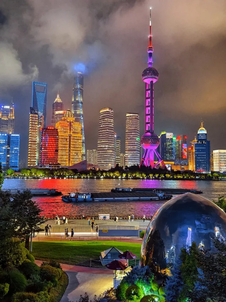 Night view of Shanghai's Pudong skyline from across the Huangpu River. The Oriental Pearl TV Tower is prominently lit in purple, alongside other illuminated skyscrapers like the Shanghai Tower and Jin Mao Tower. In the mid-ground, boats traverse the river, while the foreground features a lively waterfront promenade with people, green park areas, and a large reflective sphere sculpture.
