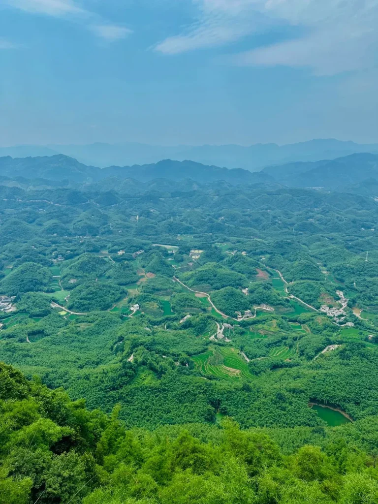Panoramic aerial view of a vast landscape of rolling green hills densely covered in bamboo forests, with winding roads, scattered small villages, and a small green lake visible in the distance under a hazy blue sky.