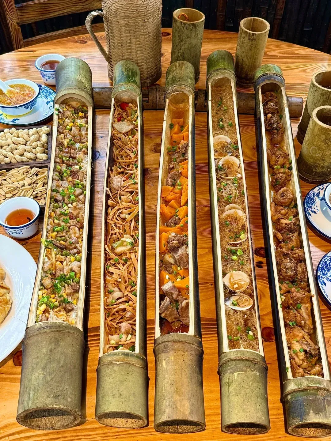 Five long bamboo tubes display a variety of steaming Sichuan dishes on a wooden table: rice with pork, corn, and green onions; stir-fried bamboo shoots with meat; pumpkin and pork ribs; glass noodles with clams; and braised chicken. A bamboo pitcher, small bamboo cups, a bowl of soup, peanuts, sunflower seeds, and tea are also visible.