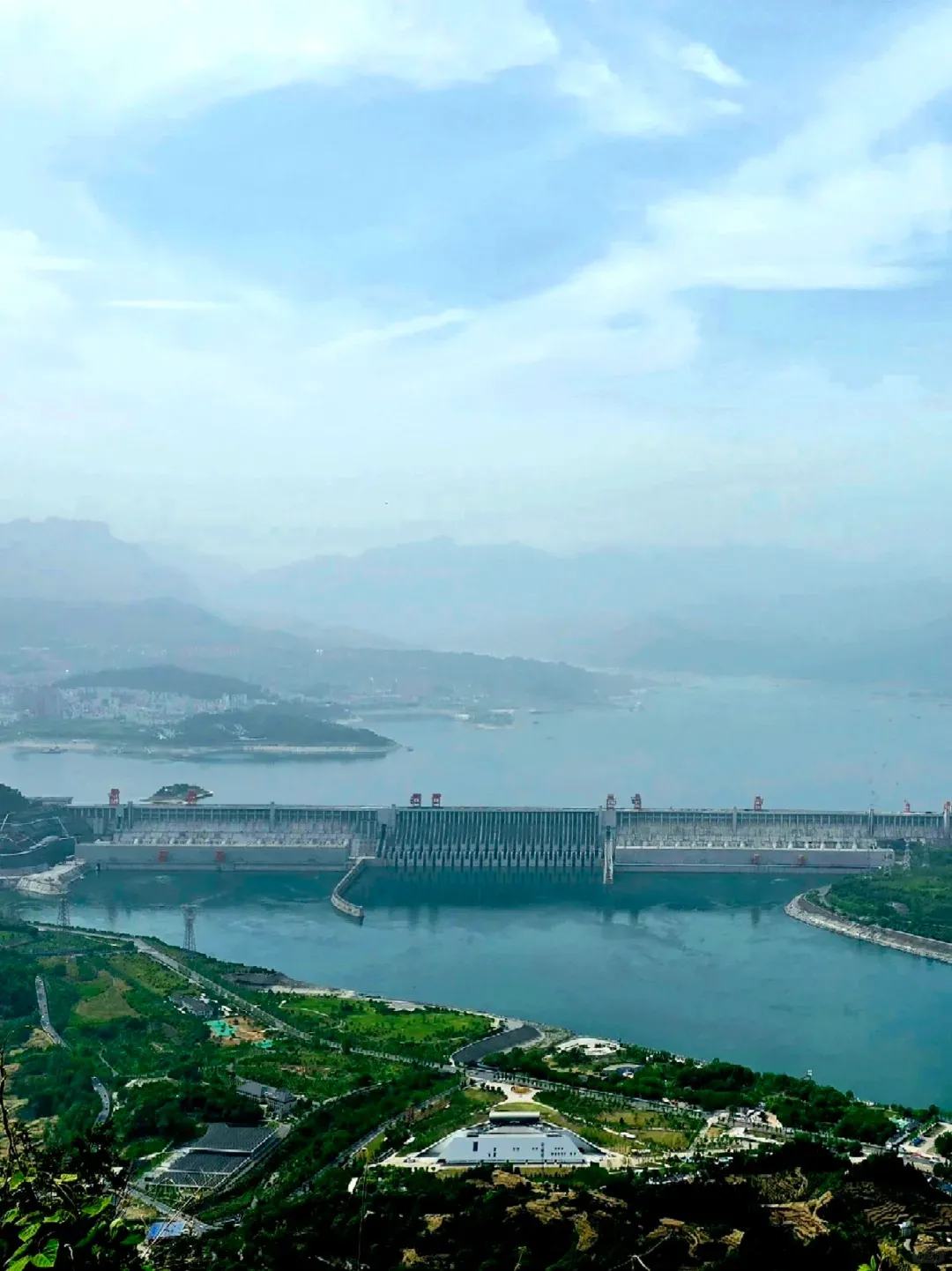 An expansive aerial view of the Three Gorges Dam spanning the wide Yangtze River in China. The massive concrete dam structure, featuring numerous gates, holds back a vast reservoir. Green, rolling hills and distant mountains, partially obscured by haze, flank the river. In the foreground, a developed area with green landscaping, roads, and a large white modern building complex is visible.