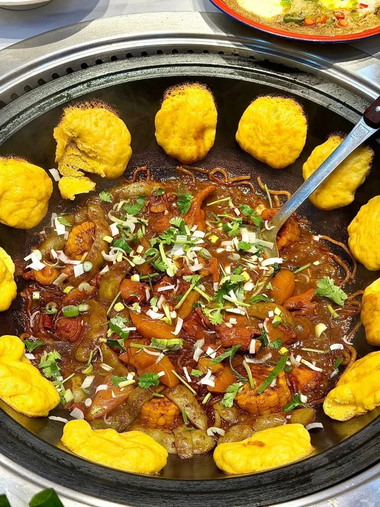 A large, circular cast iron pot filled with a rich, dark stew containing pieces of meat, vegetables like corn and green peppers, and noodles. Around the perimeter of the pot are golden yellow, steamed buns. The stew is garnished with chopped green onions and cilantro.