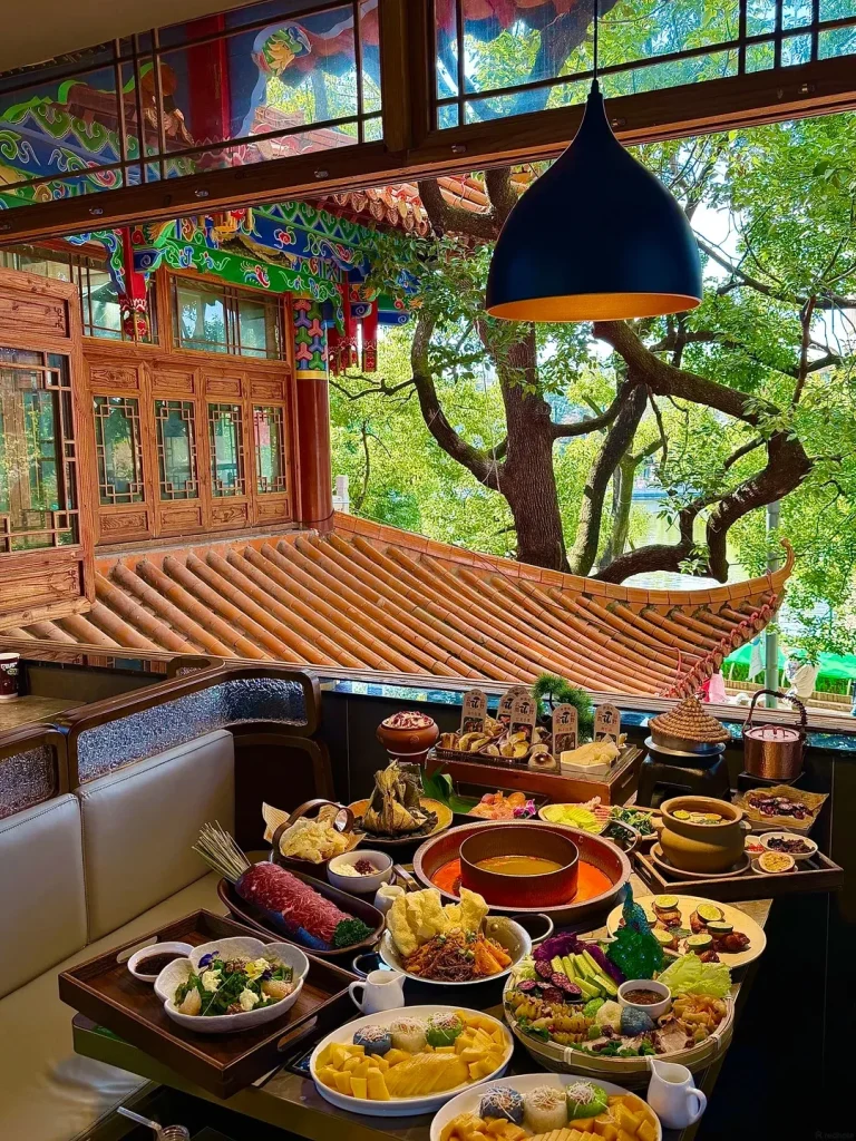 A wide shot of a lavish spread of Chinese dishes on a table in a traditional restaurant with intricate wooden carvings, painted eaves, and a tiled roof visible outside, overlooking lush green trees. A hot pot is central to the table, surrounded by various platters of meat, vegetables, fruits, and prepared foods, including sliced mango and colorful rice desserts. A dark pendant lamp hangs in the foreground.