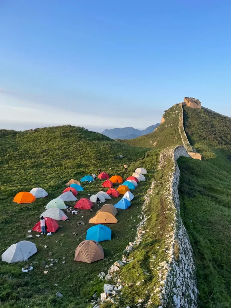 Numerous colorful camping tents are set up on a green grassy hillside next to a long, winding section of the Great Wall. The ancient stone wall extends up a steep hill to a stone watchtower on the summit, all under a clear blue sky with distant mountains in the background. A few people are visible near the tents.