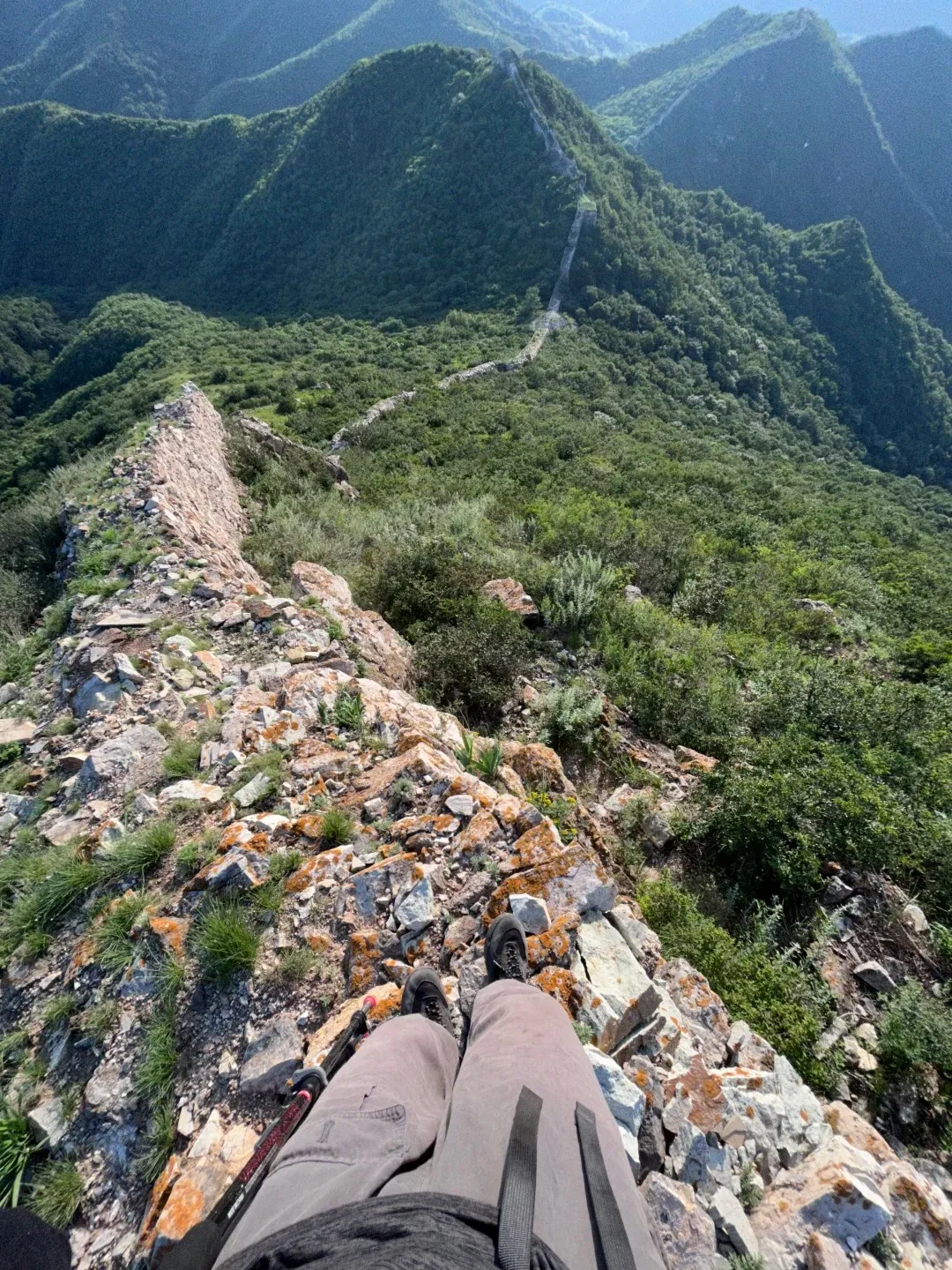 First-person view of a hiker's legs and dark boots resting on the rocky, crumbling edge of an unrestored Great Wall section, overlooking expansive green, forested mountains stretching into the hazy distance.