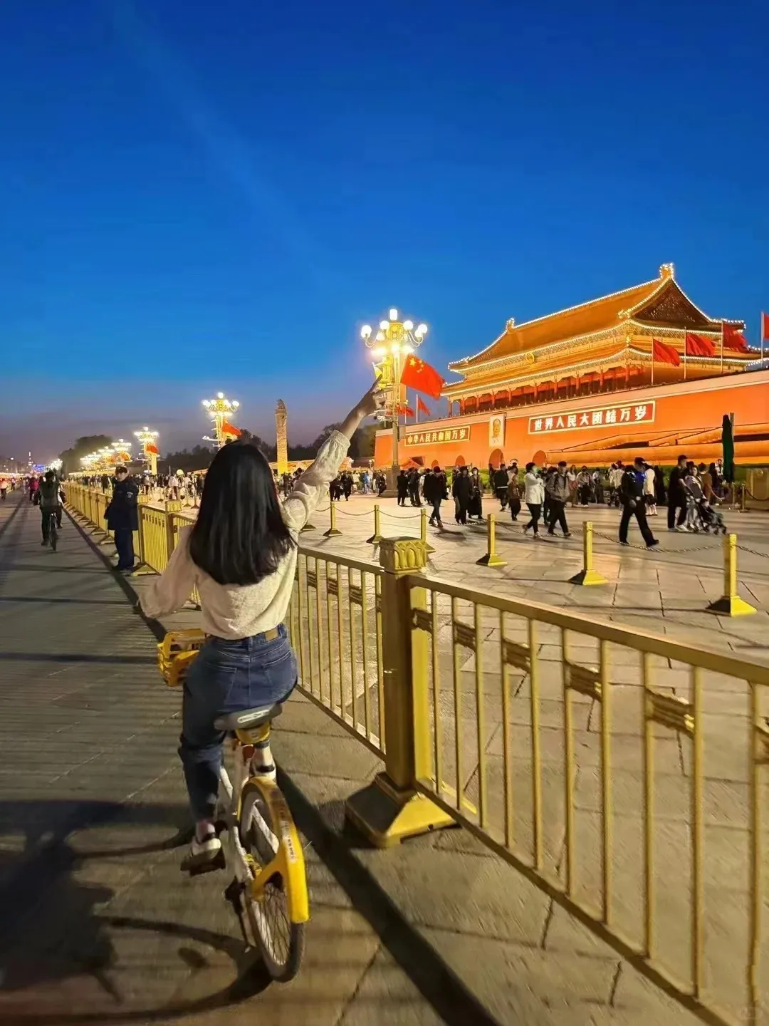 A woman riding a yellow shared bicycle, viewed from behind, points her right arm towards a Chinese flag attached to a lamppost. In the distance, the brightly lit red Tiananmen Gate stands against a deep blue sky, with many people walking in front of it and yellow barriers lining the path.