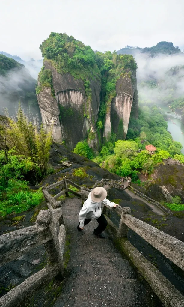 A person in a light jacket and straw hat descends a steep, mossy stone staircase with concrete railings. In the background, towering, mist-shrouded green cliffs rise above a winding river with several small boats, and a red-roofed traditional pavilion is visible among the trees.