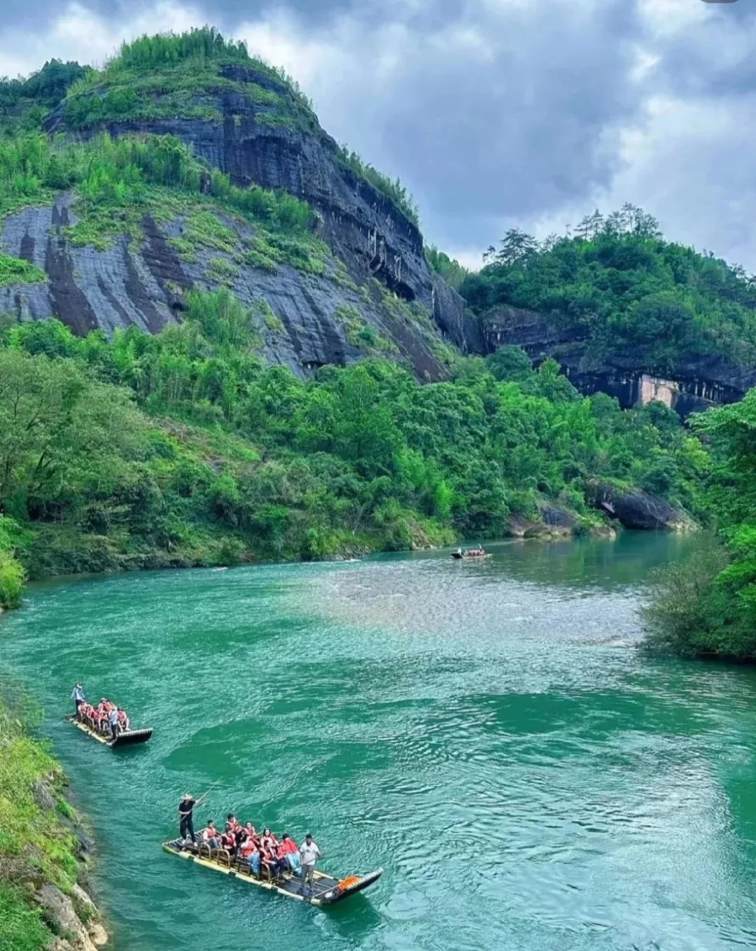 Two long bamboo rafts carrying groups of people wearing orange life vests navigate a wide river with bright turquoise-green water. The river is surrounded by tall, steep mountains covered densely with dark green trees and patches of exposed dark grey rock. The sky above is cloudy.