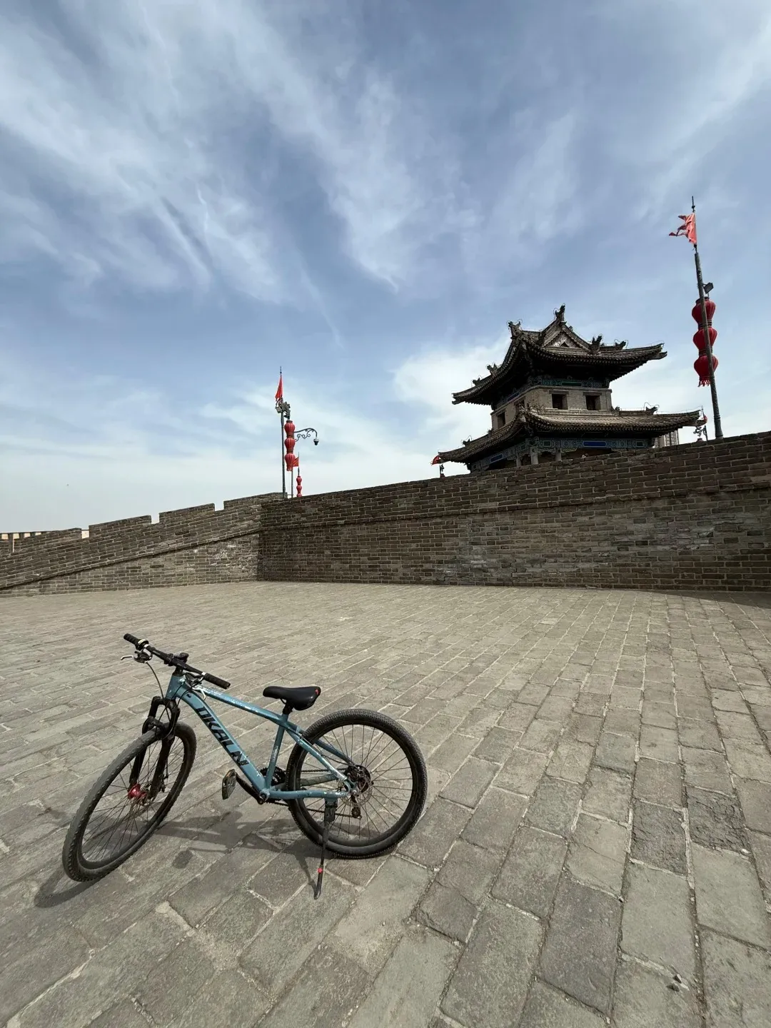 A light blue mountain bicycle with 'OKALN' on its frame leans on a kickstand on a grey brick path. In the background is an ancient grey brick city wall leading to a multi-tiered traditional Chinese watchtower with red flags and lanterns on poles, under a blue sky with wispy clouds.