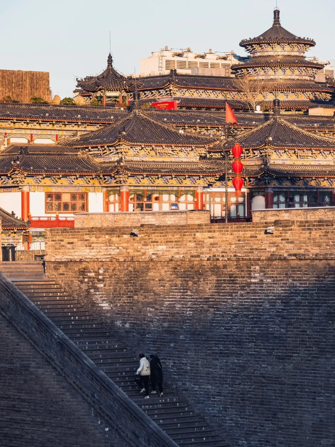A large, dark gray stone staircase leads up a massive brick wall. Two people wearing coats walk up the stairs. Above the wall, a complex of traditional Chinese buildings with multiple dark-tiled, ornate roofs, red pillars, and colorful eaves are visible. A prominent multi-tiered pagoda stands on the right, alongside red lanterns and a red flag. Modern white buildings are faintly visible in the far background. The upper section of the wall and buildings are bathed in warm, golden light.