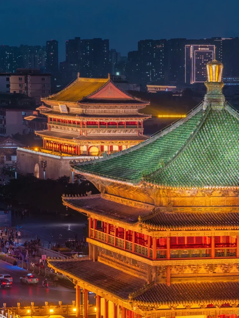 Night aerial view of Xi'an's illuminated Bell Tower (green roof in foreground) and Drum Tower (golden roof in background), with city lights and street traffic below.