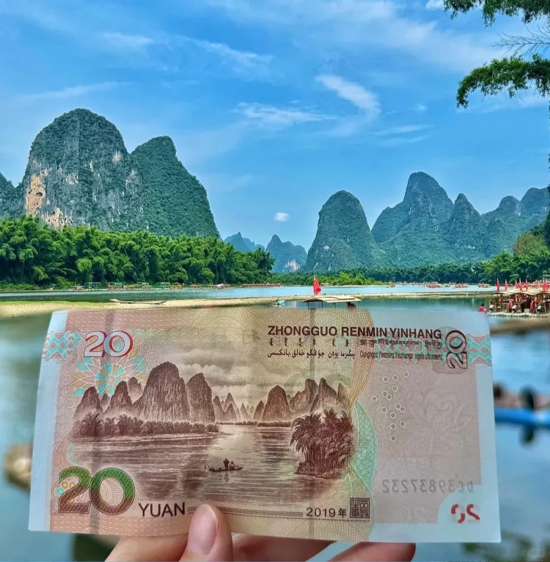 A hand holds a 20 Yuan Chinese banknote, its printed image of karst mountains along the Li River aligning with the actual lush green karst mountains in the background. Several traditional bamboo rafts are visible on the river under a clear blue sky near Xingping.