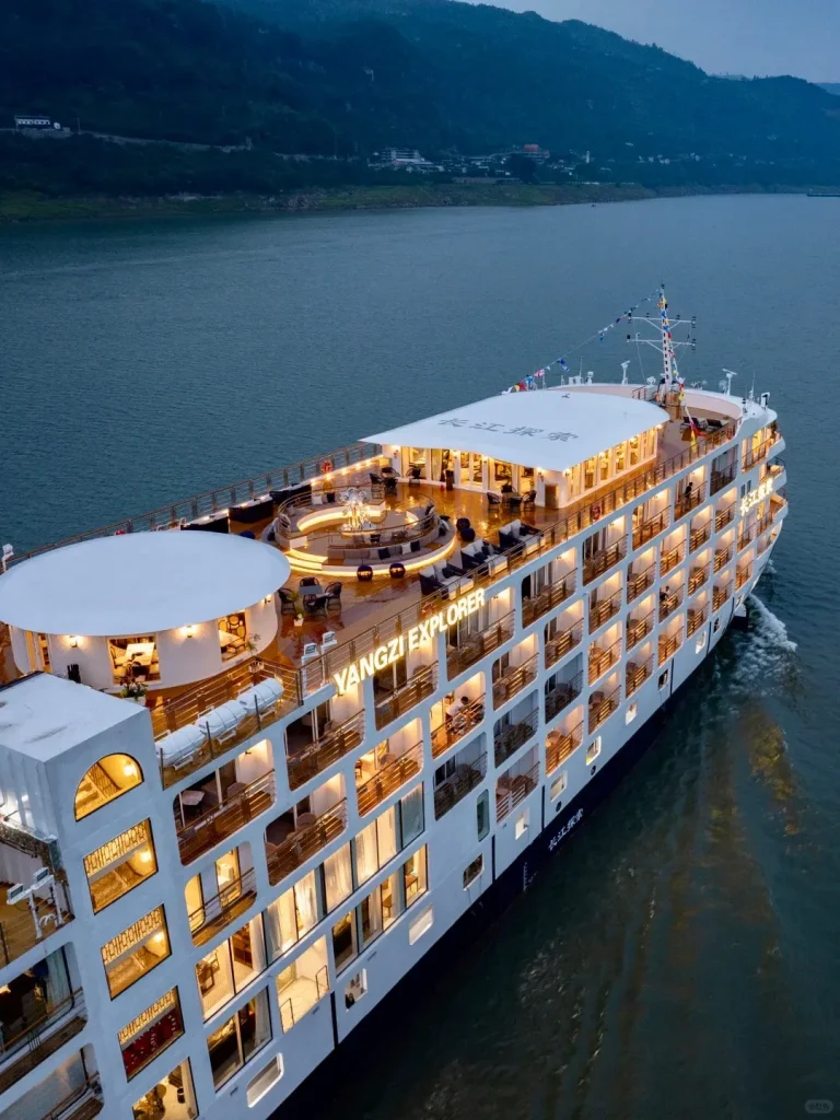 Aerial view of the multi-deck white Yangzi Explorer luxury cruise ship sailing on a dark river at dusk, brightly lit with cabin lights and deck lighting. The ship has multiple decks with balconies, and dark green mountains line the riverbank in the background.