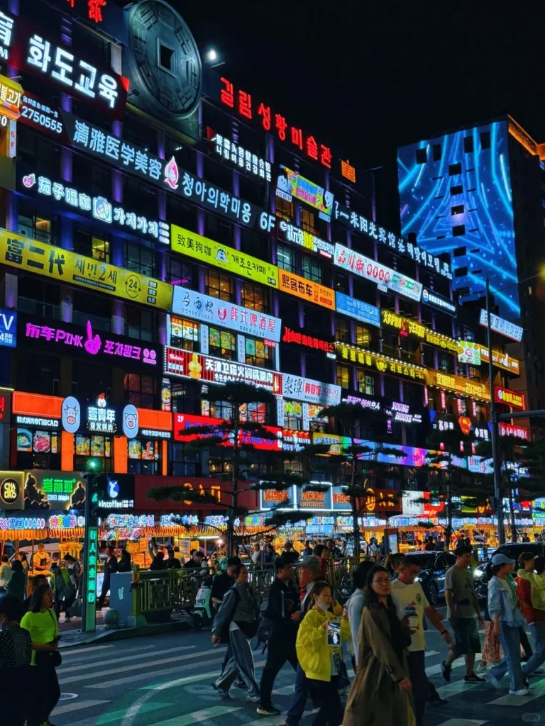A multi-story building in Yanji, China, illuminated at night with a multitude of glowing neon signs displaying both Chinese and Korean text. A large, dark, circular coin-shaped structure is mounted on the top of the building. Below, a bustling street scene shows pedestrians crossing a crosswalk under the colorful light spill from the signs, with more illuminated shops and street activity visible at ground level.