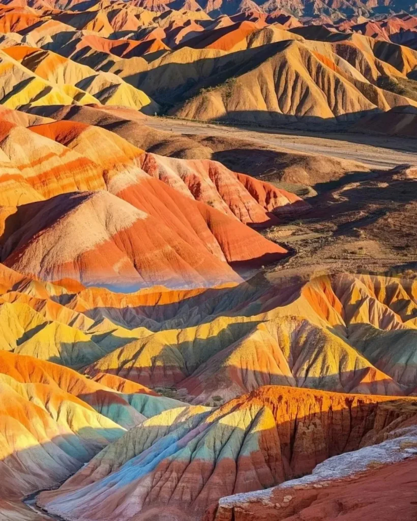 An aerial view of the Zhangye Danxia National Geopark in Gansu, China, showing numerous rolling hills and distinct sandstone ridges striped with vibrant horizontal bands of red, orange, yellow, green, and blue. Sunlight illuminates the peaks, casting long shadows into the valleys, revealing a winding road through the landscape.
