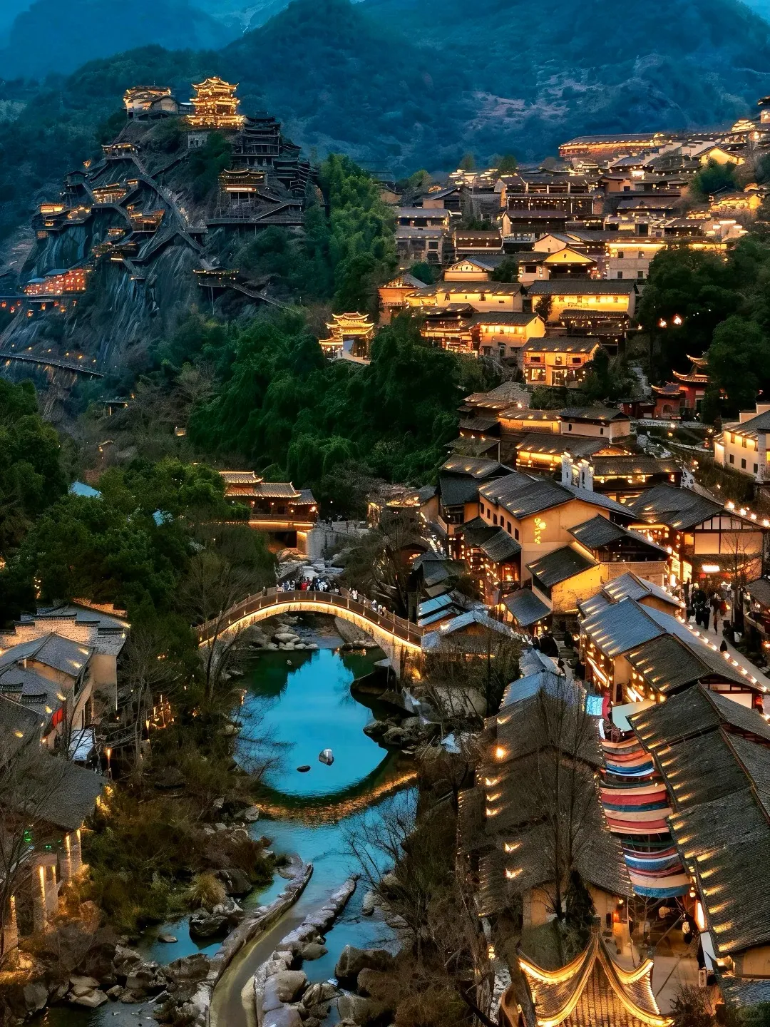 An aerial view of Zhenyuan Ancient Town at night. Traditional Chinese buildings with warm lights line both sides of a jade-colored river, which reflects the lights. A brightly illuminated arched bridge with people crosses the river in the center. On the left, numerous traditional buildings with tiered roofs are built into a steep, rocky, tree-covered mountain, also glowing with lights. Dark mountains are visible in the background.