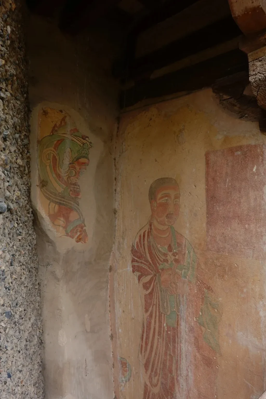 Faded ancient frescoes on a cave wall depicting a bodhisattva figure in red and green robes and a separate, larger depiction of a stylized, colorful face with green headdress and red accents. The wall has a rough texture and shows signs of age and damage. A rough stone texture is visible on the left side of the image.