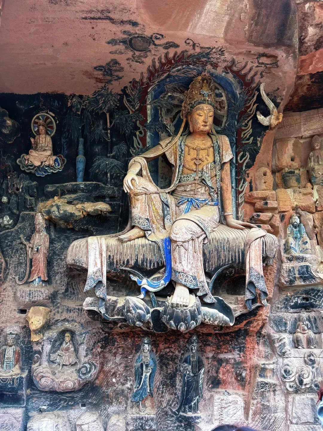 A large, seated stone sculpture of Guanyin (Purple Bamboo Guanyin) with a crown, adorned in flowing robes, resting on a lotus pedestal carved into a rock face. Smaller figures and decorative carvings surround the main sculpture within a grotto.