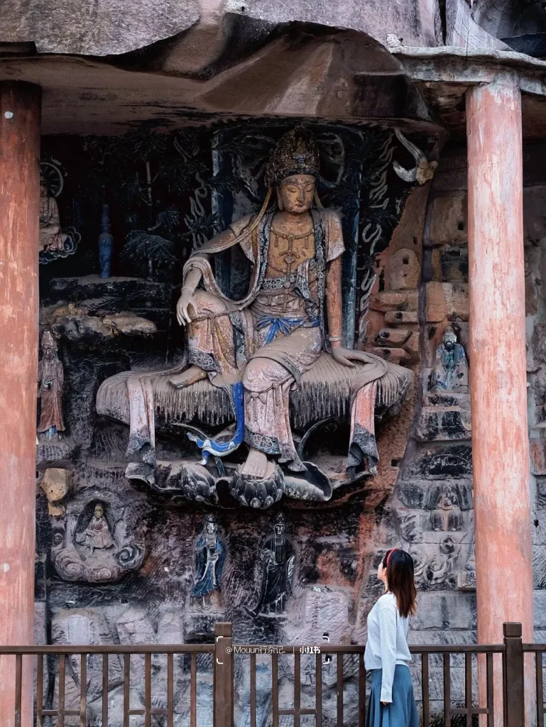 A large, ancient stone carving of Guanyin seated on a lotus throne, adorned with ornate jewelry and flowing robes, set within a cave-like structure. The statue is weathered and colorful, with intricate details in the carving of the deity and the surrounding rock formations. A young woman with a red headband stands on the right, looking up at the statue from behind a wooden railing.