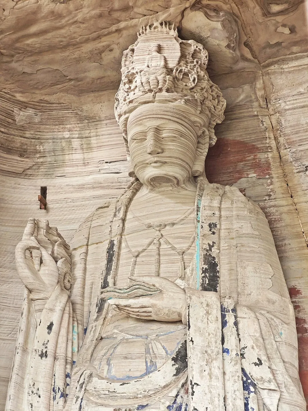 A close-up, low-angle view of a large, weathered stone carving of a serene Buddhist figure, likely a Bodhisattva, set within a rock grotto. The statue has intricate details in its headdress, facial features, and flowing robes, with visible layers of sedimentary rock forming its structure. Patches of blue and red pigment are still visible on the surface, indicating past coloration.