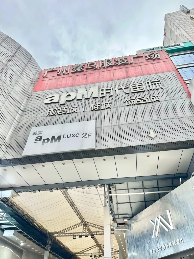 A low-angle view of a modern building facade with large signage. The top sign reads 'Guangzhou Baima Clothing City' in Chinese characters. Below it, larger white lettering says 'apm International' followed by Chinese characters for clothing, shoes, and accessories. A smaller sign on the left reads 'Korea apm Luxe 2F' with an arrow pointing right. The building has a perforated metal exterior. Below the signs is a white awning structure with exposed metal framework.