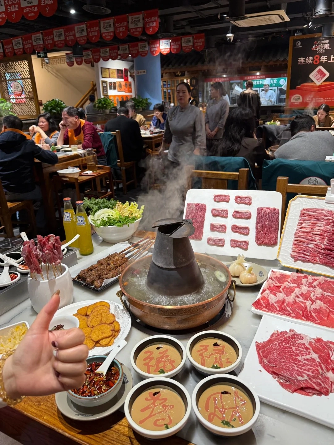 A table laden with a traditional copper pot hot pot, plates of raw sliced mutton, skewers of grilled lamb, dipping sauces, and side dishes, with people dining in the background.