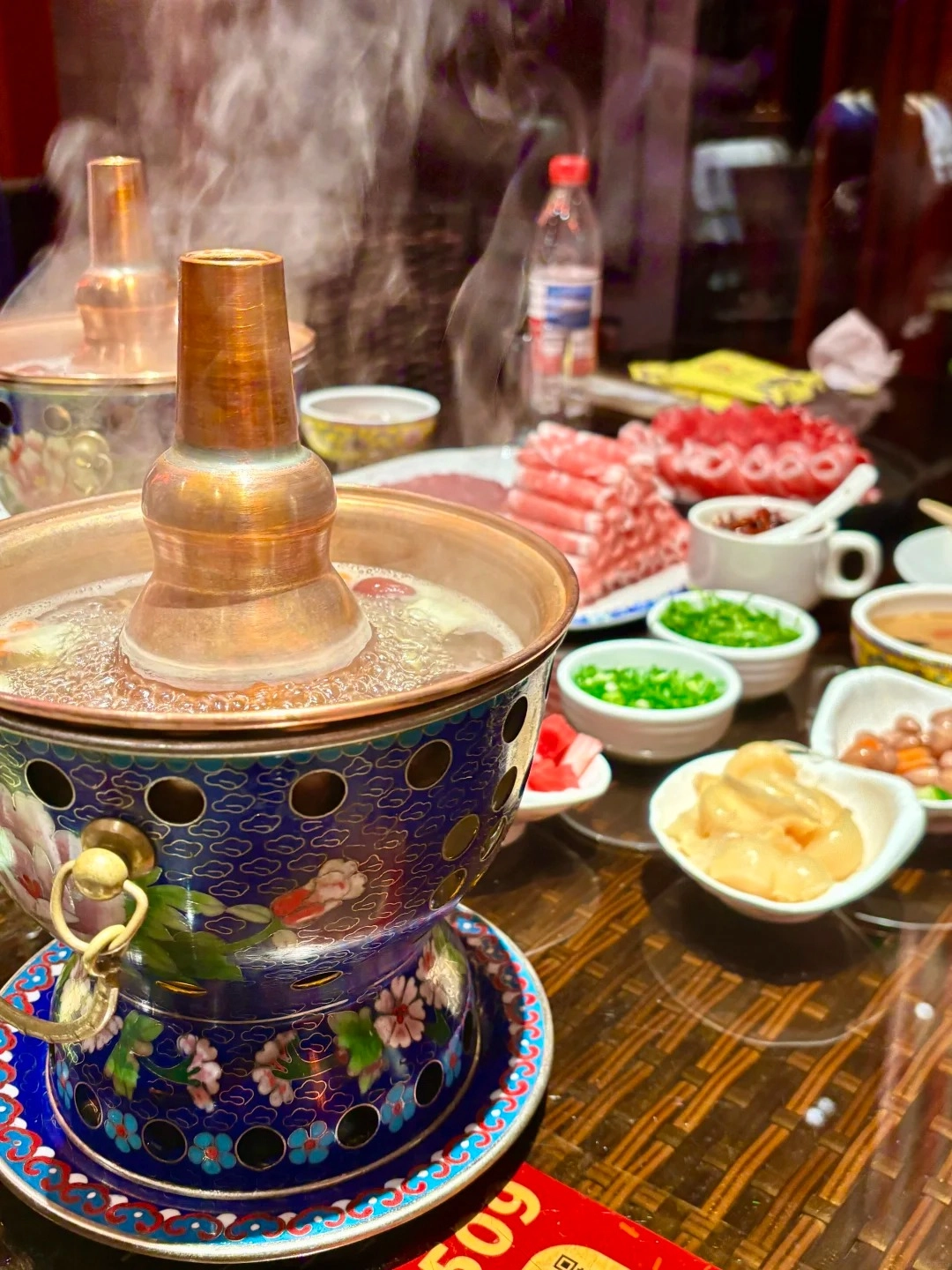 A close-up of a steaming copper hot pot with a decorative blue and floral pattern, surrounded by small bowls of ingredients including thinly sliced raw mutton, chopped green onions, and a white bowl with a red condiment. Another copper hot pot is visible in the background, along with a bottle of water.