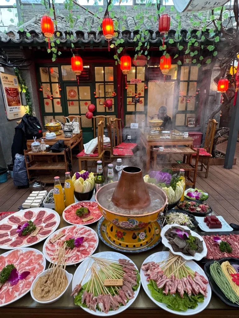 A large table laden with dishes for hot pot dining. In the center is a steaming copper pot with a decorative yellow and blue base. Around it are platters of thinly sliced raw meats, including lamb and beef, arranged in circular patterns. Several skewers of small meat pieces and a bowl of dipping sauce are also visible. Various vegetables, including napa cabbage and purple cabbage, are placed in bowls. Bottles of drinks and stacks of tofu are on the table. In the background, traditional Chinese architecture with red lanterns and green vines can be seen, with people dining at tables.