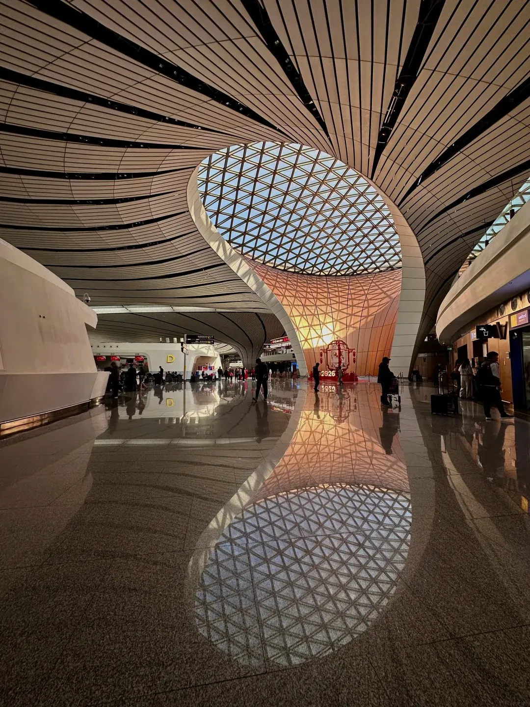 Wide-angle interior view of a modern airport terminal with a large, organically shaped skylight and curved ceiling. Sunlight streams through the skylight, casting a warm glow on the polished floor, which reflects the architectural features. People are visible in the background near check-in areas and gates.