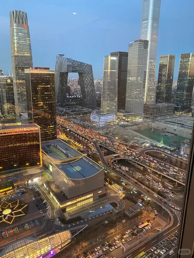 An aerial view of the Guomao Central Business District in Beijing at dusk. Several skyscrapers, including the CCTV Headquarters with its unique loop design, are visible. A complex multi-level highway interchange is packed with traffic, with car headlights and taillights creating streaks of light. Lower buildings and a parking lot with decorative star-shaped lights are in the foreground. The sky is a dusky blue with a sliver of moon visible.