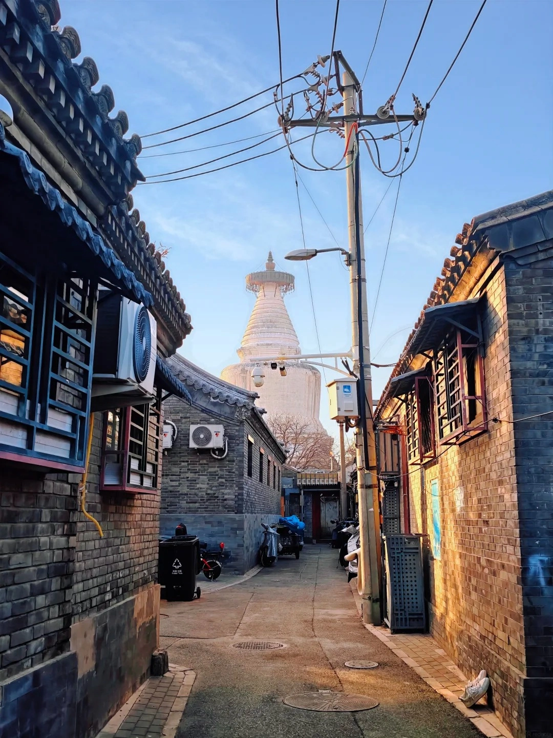 A narrow alleyway between traditional grey brick Beijing hutong buildings, leading towards a large white stupa or pagoda in the background under a clear blue sky. Power lines and a street lamp are visible in the foreground.