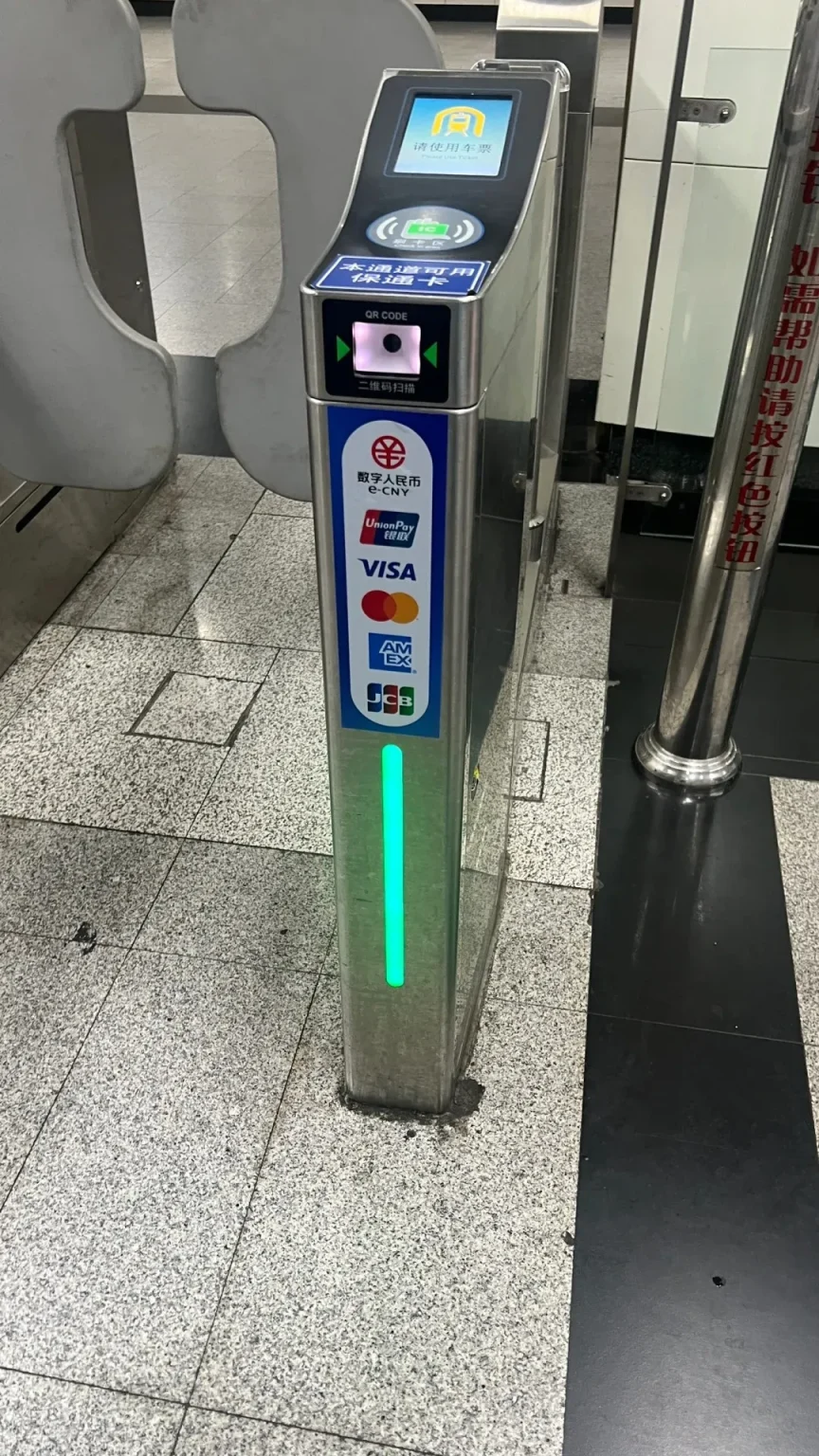 A turnstile gate in a subway station with a digital screen displaying payment options including QR codes and contactless payment logos like UnionPay, Visa, Amex, and JCB. A green light indicates the gate is operational.