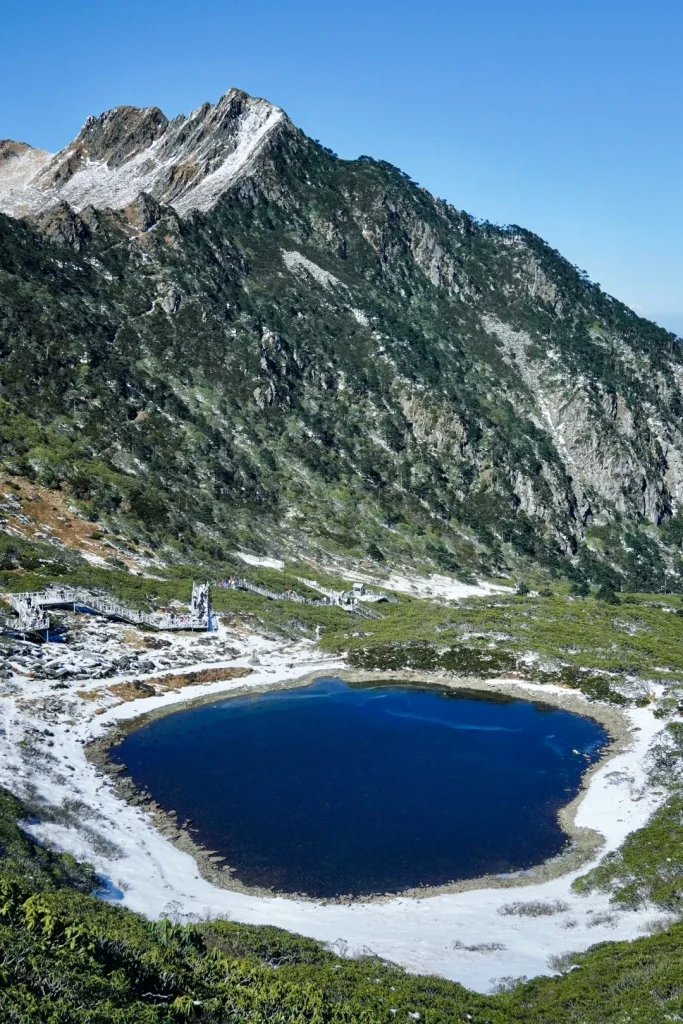 A deep blue, still alpine lake is surrounded by snow-dusted green vegetation and rocky shores. In the background, a steep, tree-covered mountain slopes upward, with patches of snow near its rocky peak under a clear blue sky. A wooden boardwalk with railings winds along the left side of the lake, with several people walking on it.