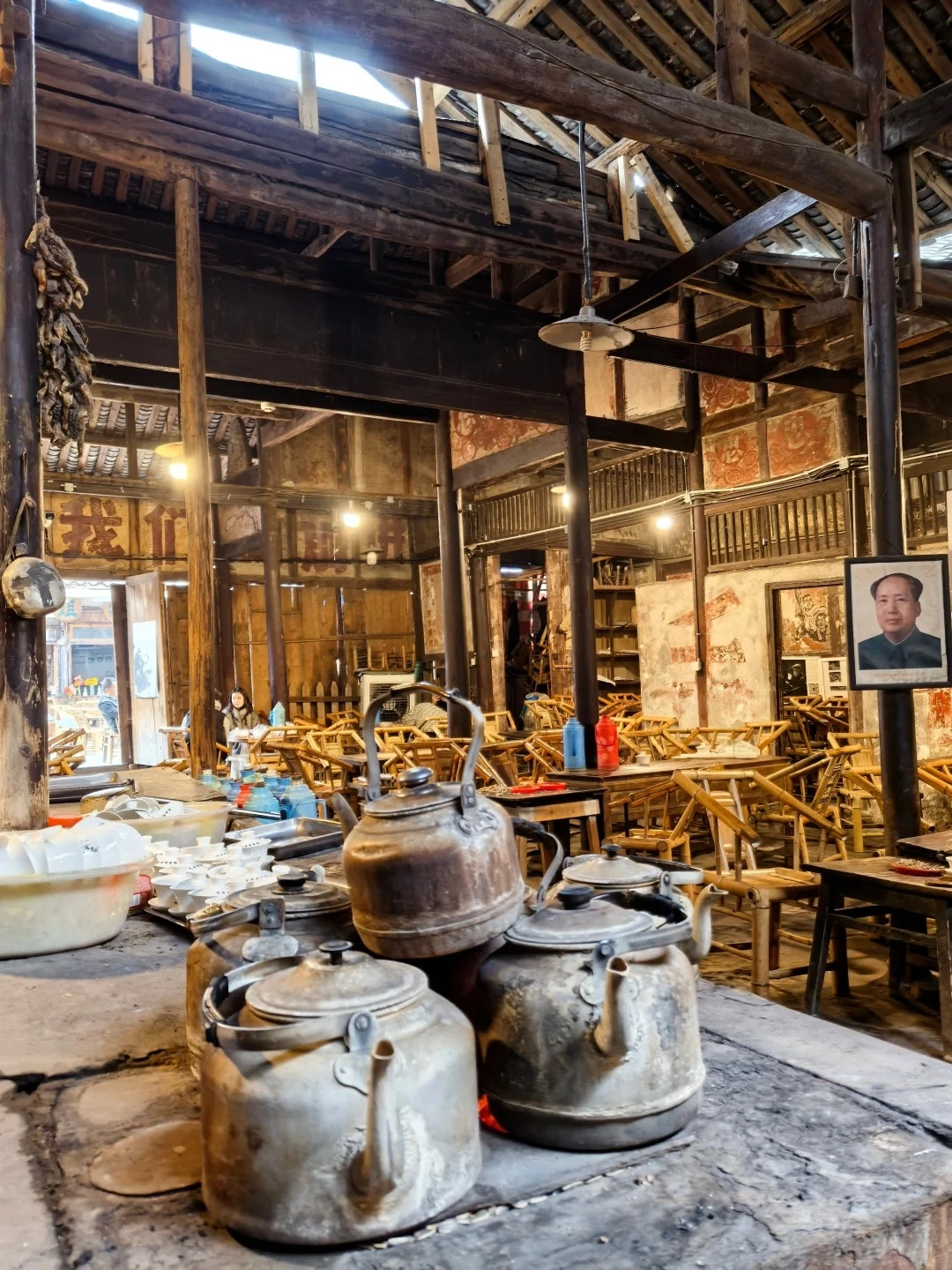A rustic interior of an old Chinese teahouse with a thatched roof and wooden beams. In the foreground, several large, weathered metal teapots are stacked on a stone surface. Behind them, a large yellow bowl and stacks of small white teacups are visible. Further back, rows of wooden tables and chairs fill the space, with some people visible in the distance.