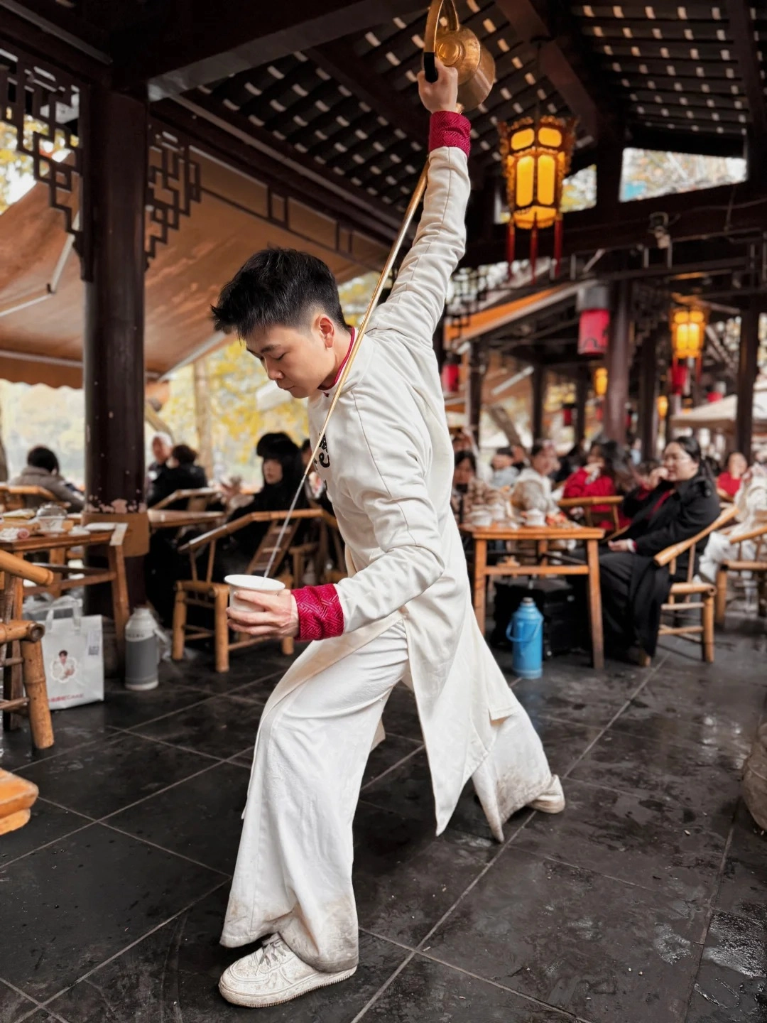 A young man in a white traditional Chinese robe with red trim on the cuffs performs a tea ceremony, holding a long metal straw with a golden bowl at the end in his right hand and a white tea cup in his left. He is standing on a dark tiled floor inside a traditional Chinese teahouse with wooden beams, decorative eaves, and glowing lanterns. Other people are visible sitting at tables in the background.