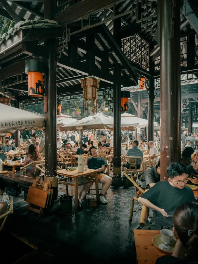 An open-air teahouse with a dark wooden roof supported by large wooden pillars. Many people are seated at bamboo tables and chairs, some eating and some drinking. In the foreground, a man in a dark t-shirt and yellow shorts leans back in his chair at a table with a bottle and a cup. Orange lanterns hang from the ceiling. The floor is wet and reflective.